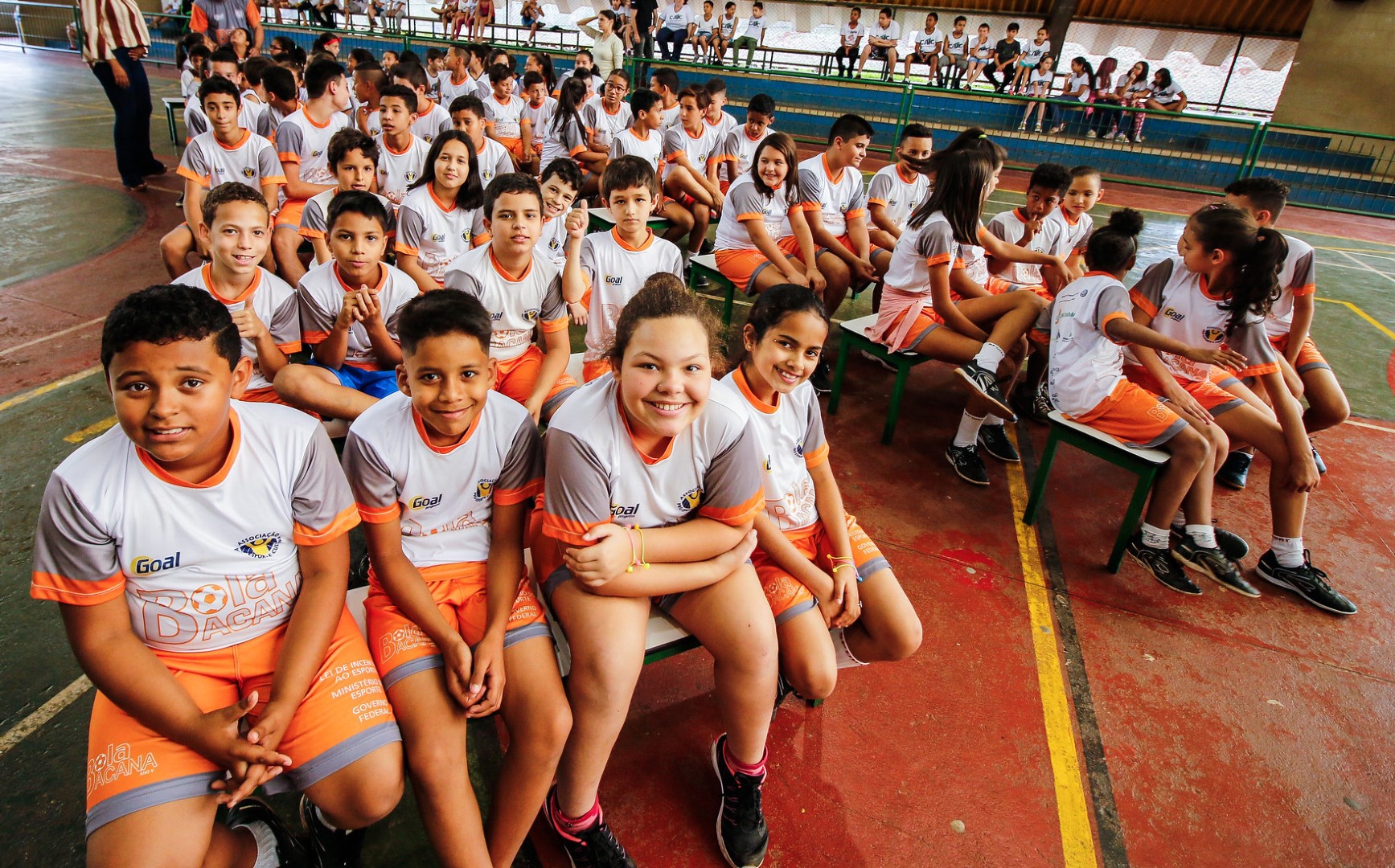 Alunos e alunas do projeto Bola Bacana vestindo uniformes brancos e cor de laranja sentados em bancos.