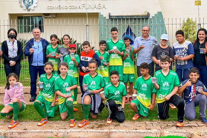 Alunos e alunas do projeto Bola Bacana reunidos vestindo uniforme verde, junto de alguns adultos em pé.