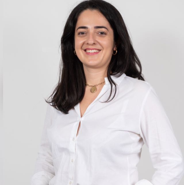 Mulher branca de cabelos pretos vestindo camisa branca e sorrindo para a foto.