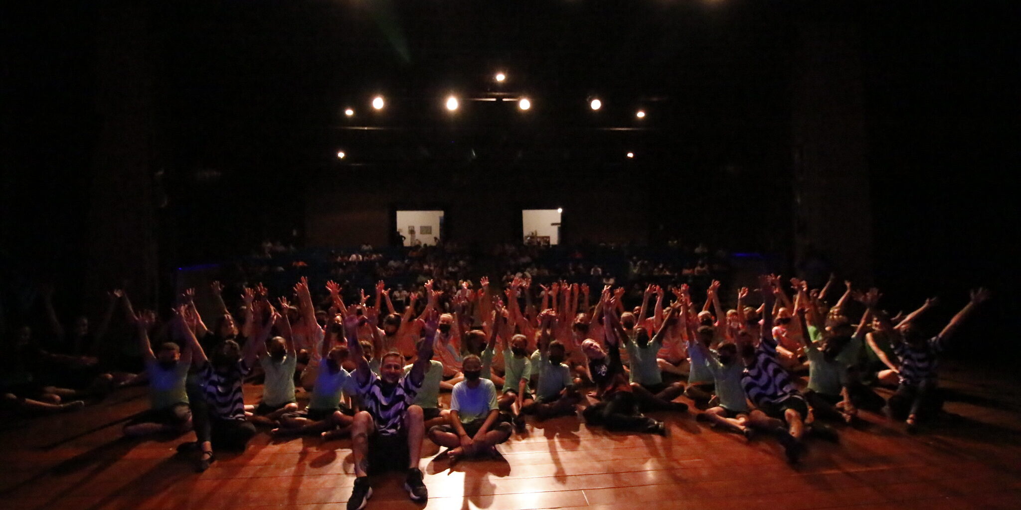 Alunos e alunas do projeto Mudança sentados em cima do palco com os braços para cima e sorrindo.