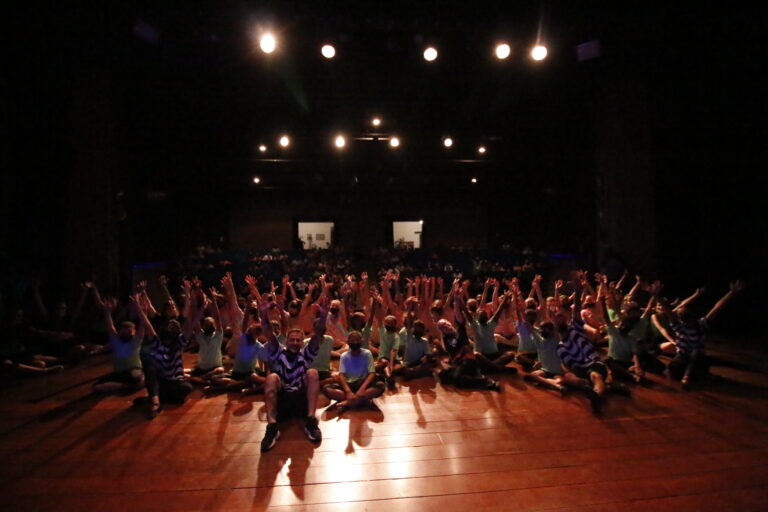 Alunos e alunas do projeto Mudança sentados em cima do palco com os braços para cima e sorrindo.